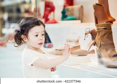 Closeup portrait of cute adorable sad upset white Caucasian toddler girl child with dark brown eyes and curly pig-tails hair in white light dress tshirt in mall looking at shoes in shop store window