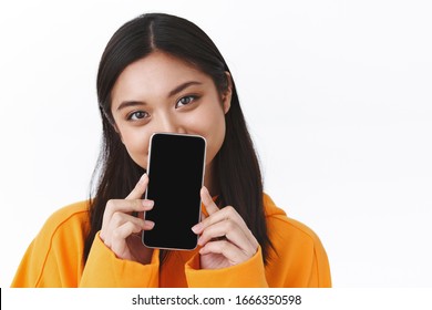 Close-up Portrait Of Cute Asian Girl In Orange Hoodie, Hiding Face Behind Smartphone, Showing Mobile Phone Display, Smiling With Eyes, Recommend Application, App Promo, White Background