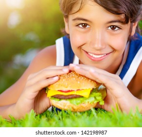 Closeup Portrait Of Cute Happy Boy Eating Big Tasty Fatty Burger Outdoors, Lying Down On Green Field And Enjoying Sandwich With Cheese, Meat And Vegetables In Sunny Day 