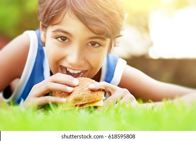 Closeup Portrait Of A Cute Happy Teen Boy Lying Down On Green Grass And Eating Delicious Burger, Enjoying Tasty Lunch Outdoors, Happy Time At Summer Camp