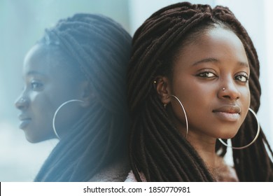 A close-up portrait of a cute young black woman with dreadlocks and huge earrings, piercing in her nose, she is leaning against a glass wall outdoors which fully reflects her and looking at the camera