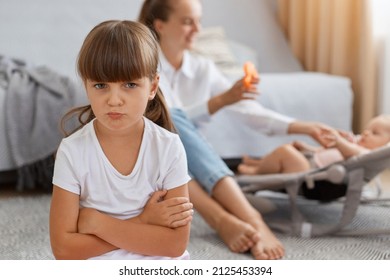 Closeup portrait of dark haired little girl wearing white t shirt posing with pout lips and unhappy expression, feels jealous mother, mommy playing with little newborn baby and don't pay attention.