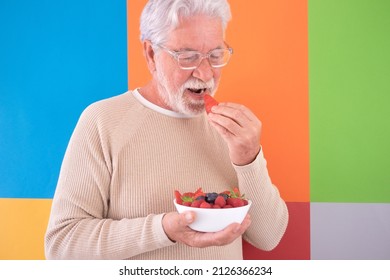 Close-up portrait of elderly mature man holding a bowl of fresh berries while biting into a strawberry. Portrait of handsome senior bearded enjoying stay fit standing against colorful background