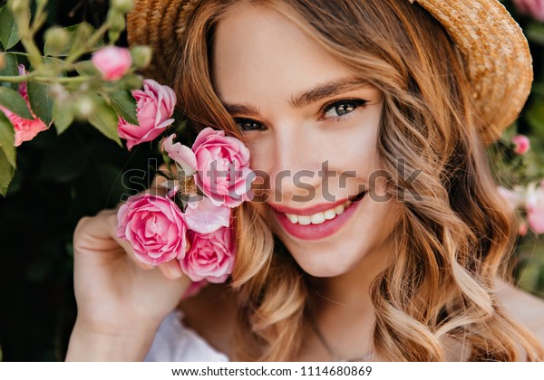 輝く目が花をポーズにしている魅惑的な女の子の接写 ピンクのバラを持つ帽子をかぶった華やかな金髪の女性が微笑みます の写真素材 今すぐ編集