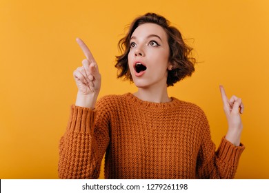 Close-up portrait of enthusiastic girl with suprised face expression looking around. Studio shot of lovable curly lady isolated on orange background.