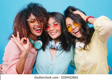 Close-up Portrait Of Excited Three Girls Laughing On Blue Background During Meeting. Indoor Photo Of Good-looking Ladies In Colorful Sunglasses Enjoying Free Time Together.