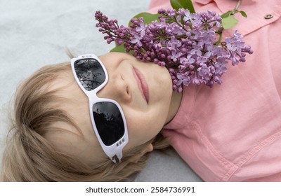 Close-up portrait of face of cute happy boy 7-9 years old in sunglasses and lilac flower, enjoys good mood and flower aroma. happy childhood. View from above