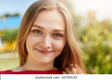 Close-up portrait of fair-haired woman with green alluring eyes, freckles smiling having dimples on cheeks looking happily directly into camera isolated over green background standing outdoors