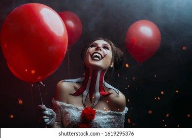 Close-up Portrait Female Clown Woman Scary Crazy Smiling Laugh Shows Sharp Teeth Predator Face, Holding Red Balloons. Backdrop Dark Gothic Room Sparks Fire Hell. Art Creative Bright Halloween Make-up