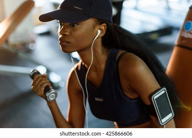 Close-up Portrait Of Fitness Instructor African American Woman Working Out With Dumbbells At The Gym And Listens Music In Earphones