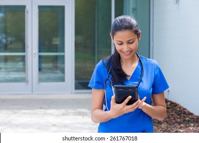 Closeup Portrait Of Friendly, Smiling Confident Female Doctor, Healthcare Professional In Blue Scrubs With Stethoscope, Analyzing Patient Data On Black Digital Tablet, Outside Hospital Background