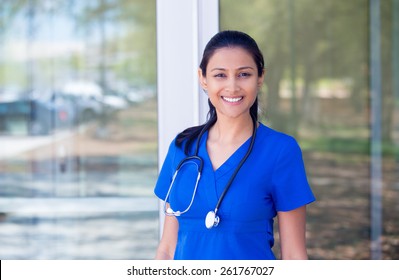 Closeup portrait of friendly, smiling confident female doctor, healthcare professional in blue scrubs with stethoscope, standing outside hospital background