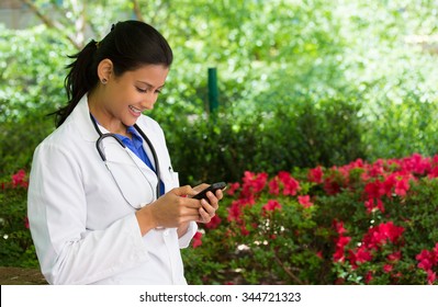 Closeup Portrait, Friendly, Young Smiling Confident Female Doctor With Stethoscope, Healthcare Professional Texting On Phone, Isolated Outside Green Trees, Red Flowers Background. Positive Emotion