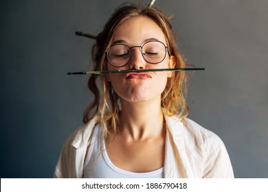 Closeup portrait of a funny beautiful female artist in eyeglasses holding with her lips a painting brush in her workshop. Pretty young woman painter making grimaces with a brush in her art studio.