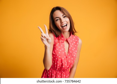 Close-up portrait of funny brunette girl showing peace gesture, looking at camera with open mouth, isolated on tellow background