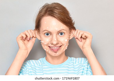 Close-up Portrait Of Funny Cheerful Girl With White Drops Of Face Cream On Her Skin, Grimacing, Making Goofy Face, Having Fun And Fooling Around, Over Gray Background. Skincare And Beauty Concept