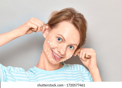 Close-up Portrait Of Funny Cheerful Girl With White Drops Of Face Cream On Her Skin, Grimacing, Making Goofy Face, Having Fun And Fooling Around, Over Gray Background. Skincare And Beauty Concept