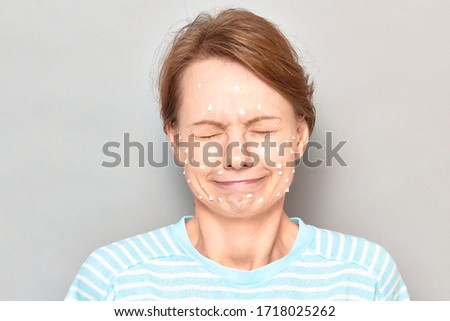 Close-up portrait of funny cheerful girl with white drops of face cream on her skin, grimacing, making goofy face, having fun and fooling around, over gray background. Skincare and beauty concept
