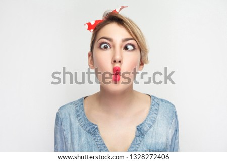 Closeup portrait of funny crazy young woman in casual blue denim shirt with makeup and red headband standing, crossed eyes with fish lips gesture. indoor studio shot, isolated on white background.