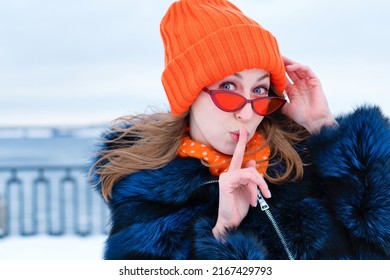 Close-up Portrait Funny Girl In Orange Beanie Hat And Blue Fur Coat On Winter Snow Background. Mysterious Young Woman Wears Stylish Orange Sunglasses Makes Gesture Secrecy Silence. Winter Walk