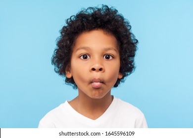 Closeup Portrait Of Funny Goofy Little Boy With Curly Hair Making Fish Face With Pout Lips And Big Eyes, Child Looking Confused With Comical Expression. Indoor Studio Shot Isolated On Blue Background