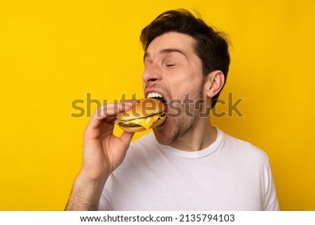 Closeup Portrait Of Funny Hungry Guy Biting Burger Eating Junk Food Holding Sandwich With Open Mouth And Closed Eyes Posing On Yellow Studio Background. Man Enjoying Big Hamburger, Bad Eating Habit