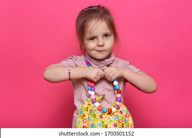 Closeup portrait of funny little girl with multicolored women's bag in hands, has jewelry, looking at camera, wearing casual shirt, posing isolated over rose studio background. Childhood concept. - Powered by Shutterstock