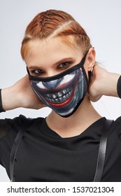 Close-up Portrait Of A Ginger Girl With Three French Braids, Posing On A Grey Background. She's Wearing Black Sweatshirt And Leather Belt. Her Face Is Hidden By Mask With Joker Smile Print.