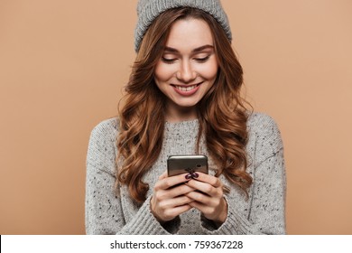 Close-up portrait of gorgeous brunette girl in woolen hat and sweater chatting on smartphone, isolated on beige background