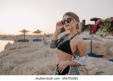 Closeup Portrait Of Gorgeous Young Girl With Slim Body Wearing Stylish Black Swimsuit, Necklace, Cardigan, Cape With Ornaments. Beach, Rocks, Sunset On Background. 