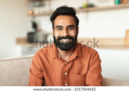 Closeup Portrait Of Handsome Cheerful Indian Man Looking And Smiling At Camera Posing At Home. Positive Millennial Arabic Young Guy With Beard Sitting On Sofa At Living Room Interior