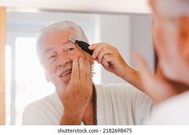 Close-up Portrait Of A Handsome Elderly Man In A Bathrobe Adjusting His Eyebrows With An Electric Razor Looking At Himself In Front Of The Mirror