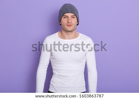 Closeup portrait of handsome magnetic man standing isolated over lilac background in studio, wearing grey hat and white sweatshirt, looking directly at camera, keeping strong fit. Cold times concept.