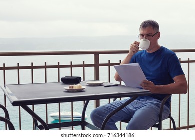 Closeup Portrait Of Handsome Man With Grey Hair Wearing Pajamas And Glasses Reading Documents Outside Balcony Isolated On A Seaside View. Man Is Having Breakfast On His Private Terrace In The Morning.