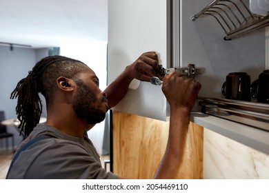 Closeup portrait handsome repairman fixing white cupboard door, using spanner. Skilled African American handyman wearing gray t-shirt in kitchen interior. - Powered by Shutterstock