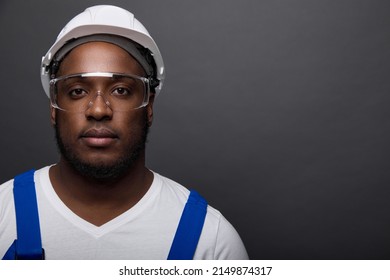 Close-up portrait of a handsome serious locksmith in a white helmet on a gray wall. In a white T-shirt and overalls with blue straps poses a young worker in construction goggles - Powered by Shutterstock