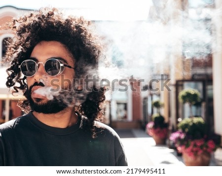 Closeup portrait of handsome smiling hipster model.Unshaven Arabian man smoking electronic cigarette. Fashion male with long curly hairstyle posing in the street. Makes smoke. Steam from vape