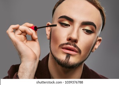 Closeup portrait of handsome young man, visagist or male makeup artist,  applying mascara, wearing gold eyeshadow, anchor beard and mustache, looking aside while holding mascara brush close to face.  - Powered by Shutterstock