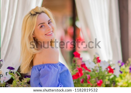 Close-up portrait of happy beauiful young woman with blond hair and pink make-up smiling on the background of  flower pots. Summer portrait of a girl