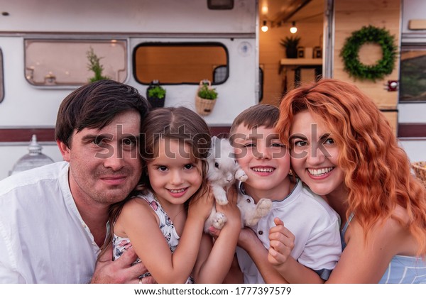 Close-up portrait of a happy family with a white\
fluffy mini rabbit near a trailer. People are looking at the\
camera, smiling cheerfully. Concept of easy travel for parents with\
two children and pets