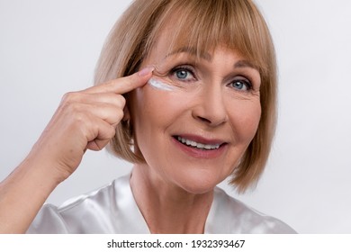 Closeup portrait of happy senior woman in dressing gown applying rejuvenating face cream on light studio background. Lovely mature lady using anti-aging cosmetic product