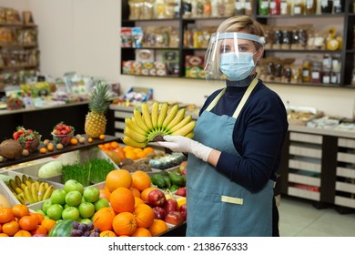 Close-up Portrait Of A Happy, Smiling Female Grocery Store Worker Wearing A Protective Mask And Gloves, Holding Bananas And Looking At The Camera.