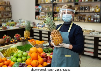 Close-up Portrait Of A Happy, Smiling Female Grocery Store Worker Wearing A Protective Mask And Gloves, Holding A Pineapple And Looking At The Camera.