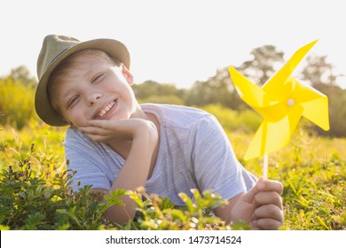 Closeup Portrait Of Happy Smiling White Kid Relaxing Happily In Sunny Sunset Meadow Outside. Boy Holding Yellow Plastic Pinwheel Toy In Hand. Horizontal Color Photography.