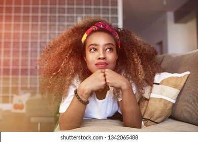 Closeup Portrait Of A Happy Young American African Girl With Long Curly Hair Relaxing And Have Fun Alone On Sofa At Modern Home.