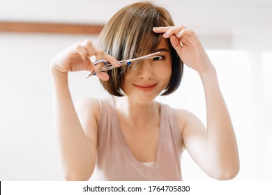 Closeup Portrait Of Happy Young Asian Woman Having Her Hair Cut With Scissors At Home. She's Stay At Home During The Coronavirus Pandemic, Self Hair Care During Quarantine.