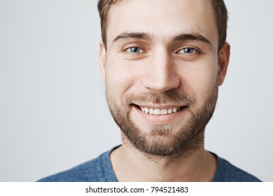 Close-up portrait of happy young male model with beard and stylish hairdo, smiles pleasantly and broadly with teeth at camera, expresses positive emotions, isolated against gray background