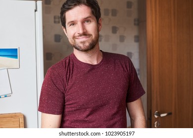 Closeup Portrait Of Happy Young Man In A T-shirt Standing Alone By The Fridge In His Kitchen In The Morning