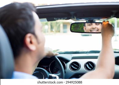 Closeup Portrait, Happy Young Man Driver Looking At Rear View Mirror Smiling, Adjusting Image Reflection, Isolated Interior Car Windshield Background. Positive Human Expression Body Language