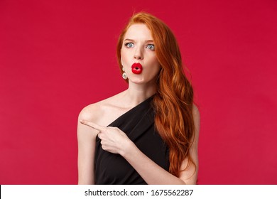 Close-up Portrait Of Impressed Excited And Speechless Attractive Redhead Woman, Wearing Black Dress, Red Lipstick And Evening Makeup. Look Startled Say Wow Pointing Left Questioned